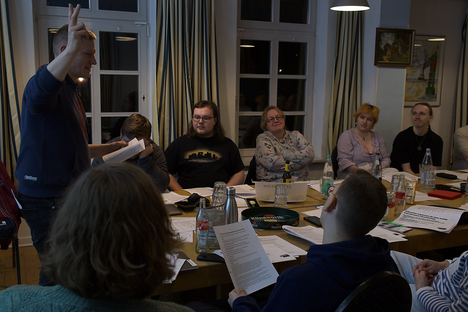 Christoph Podstawa steht vor einer Gruppe von Personen, die an Koferenztischen sitzen. Er spricht und gestikuliert. Die anderen schauen interessiert und zugewandt zu oder lesen in ausliegenden Dokumenten.